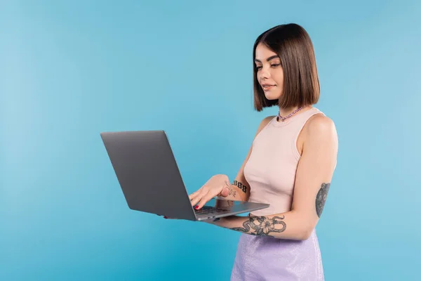 Freelancer, brunette young woman with short hair, tattoos and nose piercing using laptop on blue background, generation z, summer trends, attractive, remote work, everyday style — Stock Photo