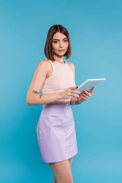 Брюнетка молода жінка з коротким волоссям, татуюваннями та носом, що пробиває цифровий планшетний ноутбук на синьому фоні, покоління z, літні тенденції, привабливі, соціальні медіа-впливові користувачі планшетів — стокове фото