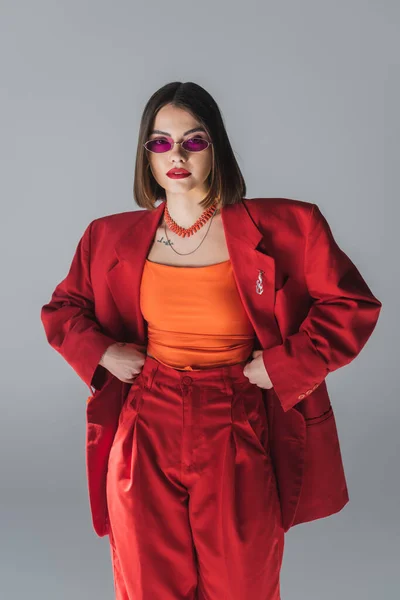 Berufskleidung, junge brünette Frau mit kurzen Haaren posiert mit rosa Sonnenbrille und rotem Anzug auf grauem Hintergrund, Generation Z, trendiges Outfit, modisches Modell, volle Länge — Stockfoto