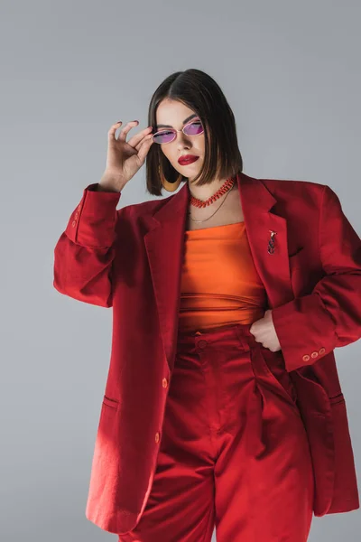 Lady in red, young brunette woman with short hair posing in pink sunglasses and red suit on grey background, generation z, trendy outfit, fashion model, professional attire — Stock Photo