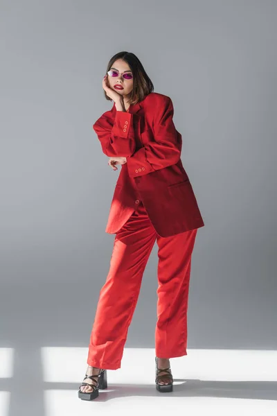 Леди в красном, молодая брюнетка женщина с короткими волосами позирует в розовых солнцезащитных очках и красный костюм на сером фоне, поколение z, модный наряд, модная модель, исполнительный стиль — стоковое фото