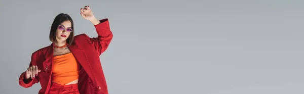 Moda gen z, joven morena con el pelo corto posando en gafas de sol de color rosa y traje rojo sobre fondo gris, generación z, traje de moda, modelo de moda, estilo ejecutivo, bandera - foto de stock