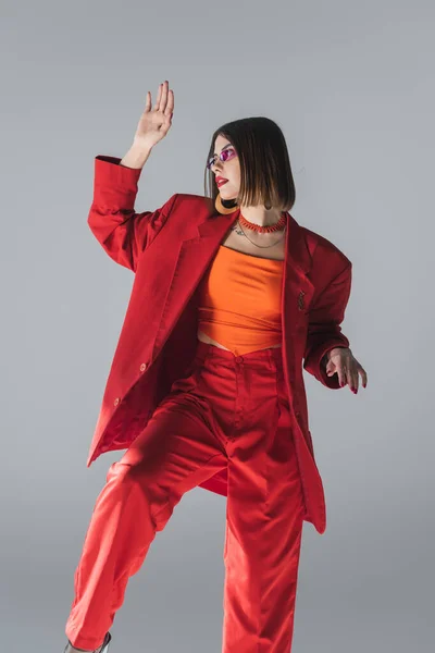 Lady in red, young brunette woman with short hair posing in pink sunglasses and red suit and gesturing on grey background, generation z, trendy outfit, fashionable model, professional attire — Stock Photo