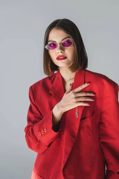 Contemporary fashion, young brunette woman with short hair posing in pink sunglasses and red suit on grey background, generation z, trendy outfit, fashionable model, professional attire — Stock Photo