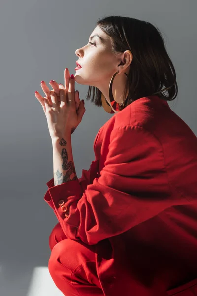 Lady in red, young and tattooed woman with short hair posing in suit on grey background, generation z, trendy outfit, fashionable model, professional attire, executive style, side view — Stock Photo