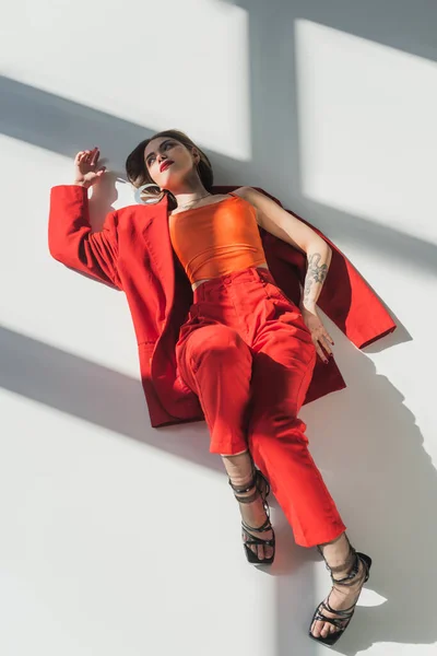 Вид сверху, молодая татуированная женщина с короткими волосами, лежащая в красном костюме на сером фоне, поколение z, модная модель, профессиональный наряд, корпоративная мода, туфли на каблуках, дама в красном — стоковое фото