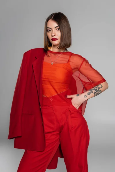 Стильный костюм, смелый макияж, молодая татуированная женщина с короткими волосами держа красный блейзер на сером фоне, поколение z, модный наряд, профессиональный наряд, исполнительный стиль — стоковое фото