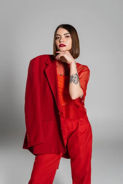 Trendy outfit, bold makeup, tattooed woman with short hair holding red blazer on grey background, generation z, professional attire, executive style, young professional — Stock Photo