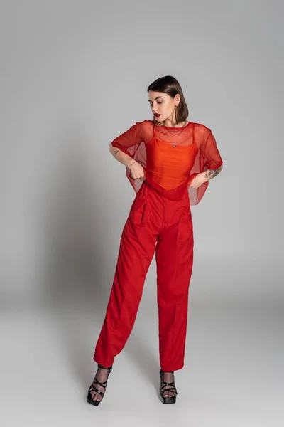 Red outfit, tattooed and brunette woman with short hair and nose piercing posing in transparent blouse and pants on grey background, modern style, generation z, fashion photography, full length — Stock Photo