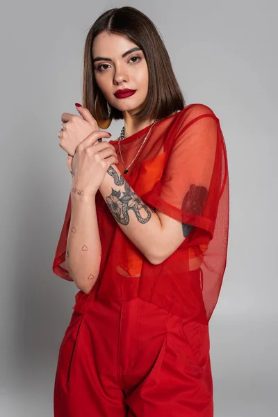 Fashionista, red outfit, tattooed and young woman with short hair and nose piercing posing in transparent blouse and pants on grey background, modern style, generation z, fashion — Stock Photo