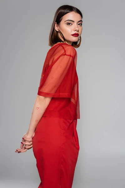 Stylish red outfit, hoop earrings, tattooed and brunette woman with short hair and nose piercing posing in transparent blouse and pants on grey background, modern style, generation z, fashion — Stock Photo