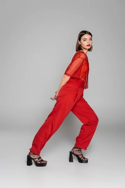 Model in red outfit, tattooed and brunette woman with short hair and nose piercing posing in transparent blouse and pants on grey background, modern style, generation z, fashion trend, full length — Stock Photo