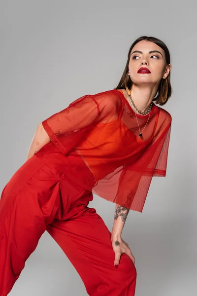 Model in red outfit, tattooed young woman with short hair and nose piercing posing in transparent blouse and pants on grey background, modern style, generation z, fashion trend — Stock Photo