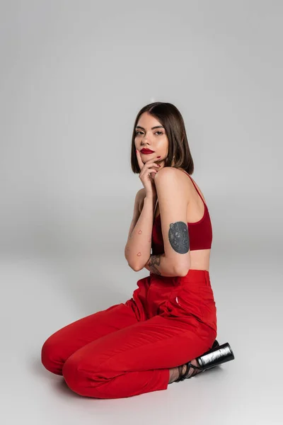 Молодая модель в красном наряде, татуированная женщина с короткими волосами и пирсингом носа позирует в красном топе и брюках, сидя на сером фоне, современный стиль, поколение z, модный тренд — стоковое фото