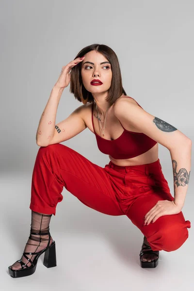 Модний тренд, молода модель в червоному вбранні, татуйована жінка з коротким волоссям і пірсингом носа позує в червоній культурі зверху і штани, сидячи на сірому фоні, сучасний стиль, покоління z, повна довжина — стокове фото