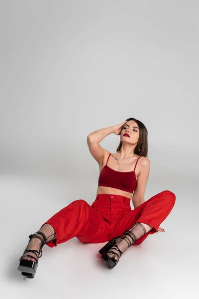 Fashion trend, chic style, young model in red outfit, tattooed woman with short hair and nose piercing posing in red crop top and pants on grey background, generation z, full length — Stock Photo