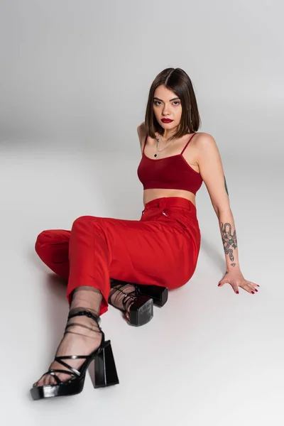 Шикарный стиль, модный наряд, молодая модель в красном наряде, татуированная женщина с короткими волосами и пирсингом носа позирует в красном топе и брюках на сером фоне, поколение z, полная длина — стоковое фото