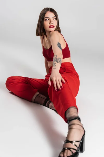 Модний одяг, молода модель в червоному вбранні, впевнена і татуйована жінка з коротким волоссям і пірсингом носа позує в червоній культурі зверху і штани на сірому фоні, покоління z, повна довжина — стокове фото