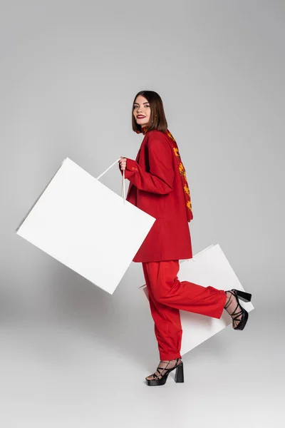 Споживання, щаслива жінка з брюнеткою коротке волосся і пірсинг носа тримає сумки для покупок і ходить на сірому фоні, сучасний модний тренд, модний одяг, червоний костюм, повна довжина — стокове фото
