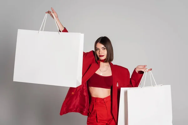 Consumismo, jovem mulher com cabelo curto morena e nariz piercing segurando sacos de compras e andando em fundo cinza, tendência de moda moderna, roupa na moda, terno vermelho — Fotografia de Stock