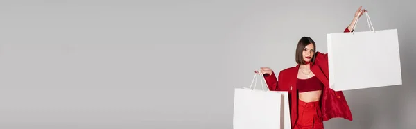 Consumismo, giovane donna con capelli corti bruna e piercing al naso che tiene borse della spesa e cammina su sfondo grigio, tendenza della moda moderna, vestito alla moda, abito rosso, banner — Foto stock