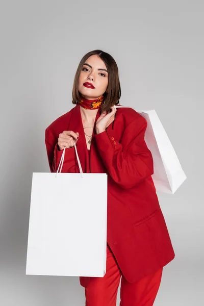 Consumerism, bold makeup, young woman with brunette short hair and nose piercing holding shopping bags and standing on grey background, youth culture, fashionable outfit, red suit — Stock Photo