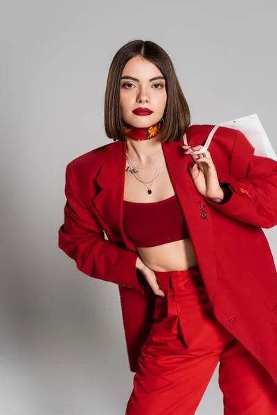 Consumerism, bold makeup, tattooed young woman with brunette short hair and nose piercing holding shopping bag and standing on grey background, youth culture, fashionable outfit, red suit — Stock Photo
