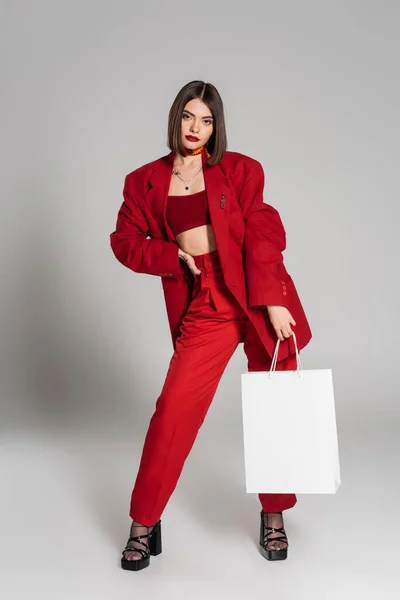 Red suit, stylish posing, tattooed young woman with brunette short hair and nose piercing holding shopping bags and standing on grey background, youth culture, fashionable outfit, consumerism — Stock Photo