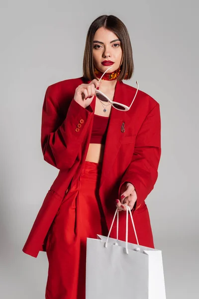 Fashionable, generation z, shocked young woman with brunette short hair and nose piercing holding sunglasses and shopping bag on grey background, youth culture, red suit, consumerism — Stock Photo