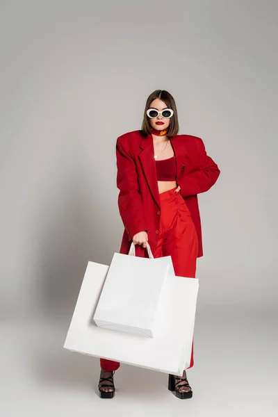 Red suit, generation z, young woman with short hair and nose piercing posing in sunglasses and holding shopping bags on grey background, youth culture, consumerism — Stock Photo
