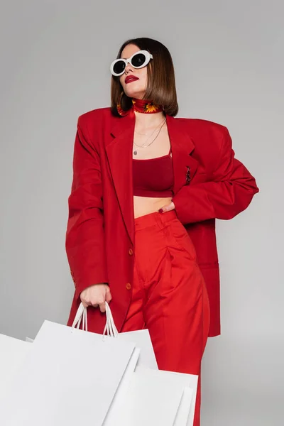 Fashionable, generation z, young woman with brunette short hair and nose piercing posing in sunglasses and holding shopping bags on grey background, lady in red, consumerism — Stock Photo