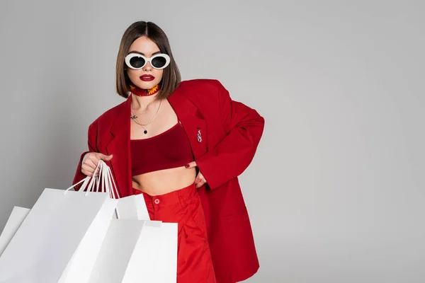Fashionable, generation z, young woman with brunette short hair and nose piercing posing in sunglasses and red suit while holding shopping bags on grey background, youth culture, consumerism — Stock Photo