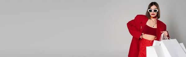 Geração z, jovem mulher com cabelo curto morena e nariz piercing posando em óculos de sol e terno vermelho enquanto segurando sacos de compras em fundo cinza, cultura juvenil, consumismo, banner — Fotografia de Stock