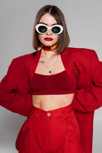Generación z, mujer joven tatuada con pelo corto y piercing en la nariz posando en gafas de sol y traje rojo sobre fondo gris, moda moderna, atuendo de moda, estilo elegante - foto de stock