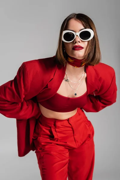 Generación z, mujer joven tatuada con pelo corto morena y piercing en la nariz posando en gafas de sol y traje rojo sobre fondo gris, moda moderna, atuendo de moda, estilo chic — Stock Photo