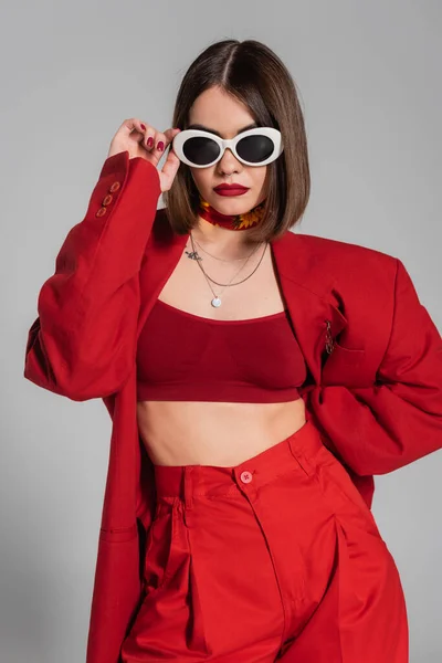 Trendiges Outfit, Generation Z, tätowierte junge Frau mit brünetten kurzen Haaren und Nasenpiercing posiert in Sonnenbrille und rotem Anzug auf grauem Hintergrund, moderne Mode, schicker Stil — Stockfoto