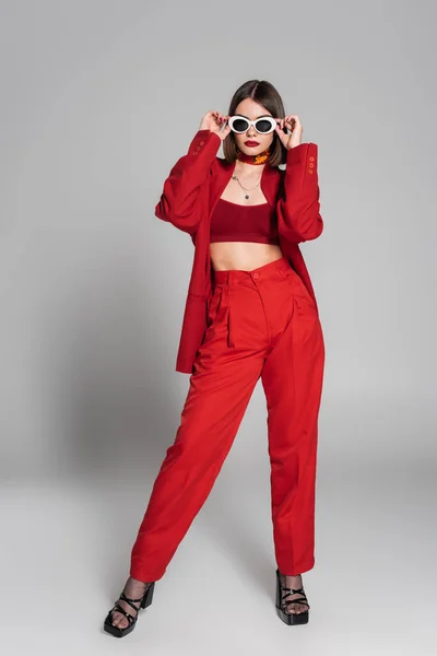 Generation z, tattooed young woman with short hair and nose piercing posing in sunglasses and red suit on grey background, modern fashion, trendy outfit, chic style, full length — Stock Photo
