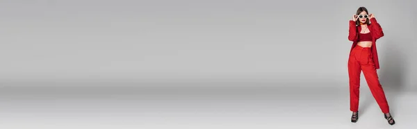 Generazione z, giovane donna tatuata con capelli corti e piercing al naso posa in occhiali da sole e abito rosso su sfondo grigio, moda moderna, abito alla moda, stile chic, lunghezza intera, banner — Foto stock