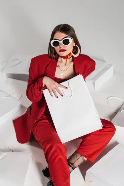 Einkaufsbummel, junge Frau mit brünetten kurzen Haaren und Piercing und Tätowierung posiert in Sonnenbrille und rotem Anzug, während sie um Einkaufstüten auf grauem Hintergrund herumsitzt, Hochblick — Stockfoto