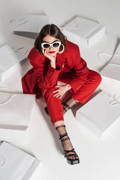 Konsumverhalten, junge Frau mit brünetten kurzen Haaren, Nasenpiercing und Tätowierung posiert in Sonnenbrille und rotem Anzug, während sie um Einkaufstüten auf grauem Hintergrund herumsitzt, Blickwinkel hoch — Stockfoto