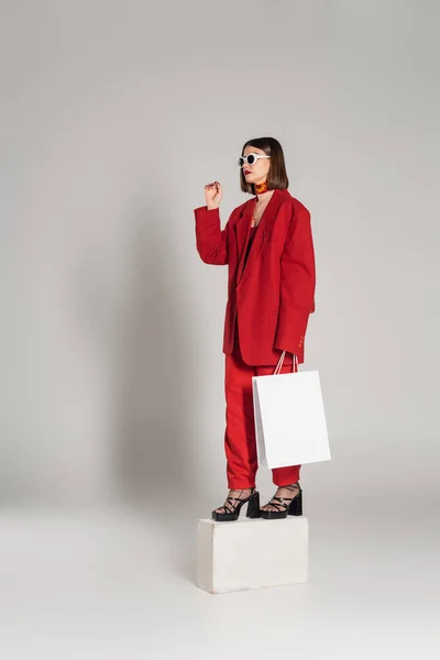 Generation z, consumerism, young woman with brunette short hair and nose piercing posing in sunglasses and red suit while holding shopping bag and standing on concrete cube on grey background — Stock Photo