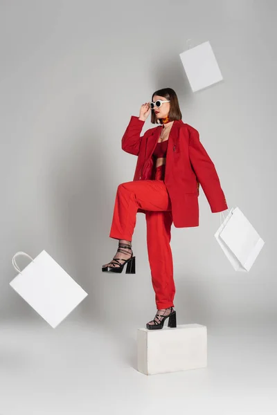 Generation z, consumerism, fashion model with brunette short hair and nose piercing posing in sunglasses and red suit while standing on concrete cube around flying shopping bags on grey background — Stock Photo