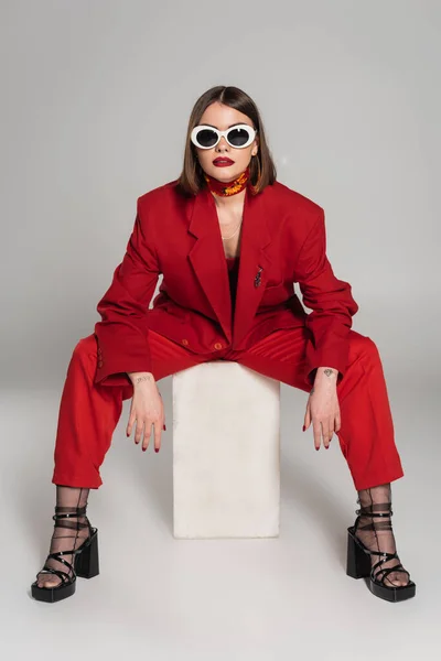 Geração z, modelo de moda com cabelo curto morena e nariz piercing posando em óculos de sol e terno vermelho enquanto sentado em cubo de concreto em fundo cinza, senhora de vermelho, fashionista — Fotografia de Stock