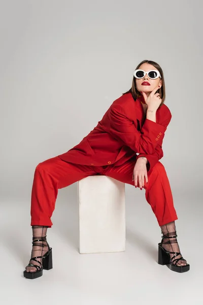 Generación z, modelo de moda con pelo corto morena y piercing en la nariz posando en gafas de sol y traje rojo mientras está sentado en cubo de hormigón sobre fondo gris, señora de rojo, mujer joven - foto de stock