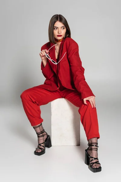 Fashion model with brunette short hair and nose piercing posing in red suit while holding sunglasses and sitting on concrete cube on grey background, lady in red, young woman, fashion trend — Stock Photo