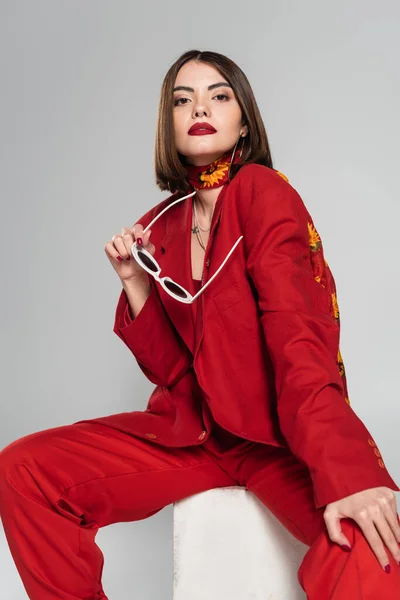 Модель моди з брюнеткою коротке волосся і пірсинг носа позує в червоному костюмі, тримаючи сонцезахисні окуляри і сидячи на бетонному кубі на сірому фоні, стильна позування, леді в червоному, молода жінка — стокове фото