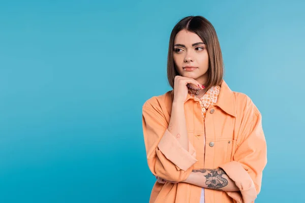 Génération z, portrait de femme concernée, jeune mannequin regardant ailleurs et pensant sur fond bleu, chemise orange, cheveux bruns courts, nez percé, tenue d'été, gen z fashion — Stock Photo