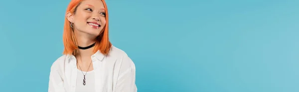 Positividad, alegre mujer joven asiática con el pelo teñido de pie en traje casual y sonriendo sobre fondo azul, camisa blanca, mirando hacia otro lado, collar de gargantilla, pelo rojo, generación z, bandera - foto de stock