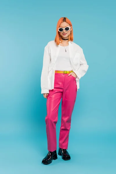 Happy face, young asian woman with dyed hair standing in casual attire and sunglasses, smiling on vibrant blue background, white shirt, choker necklace, red hair, generation z, hand in pocket — Stock Photo
