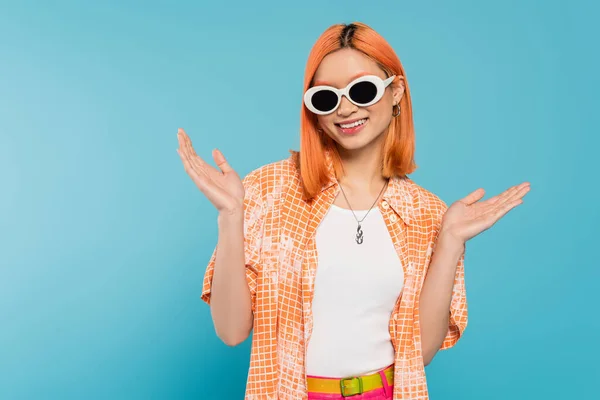 Glücklich, junge asiatische Frau mit gefärbten Haaren in lässiger Kleidung und Sonnenbrille, gestikulierend mit den Händen auf leuchtend blauem Hintergrund, orangefarbenem Hemd, Halskette, Generation z, rotes Haar — Stockfoto